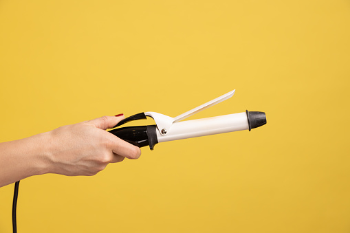 Closeup side view of woman hand holding showing iron curler, professional curling tongs, beauty care concept and hairstyling. Indoor studio shot isolated on yellow background.