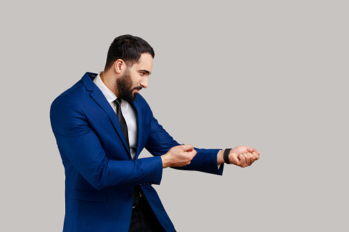 Side view of bearded man looking diligent and strong, standing in attack or pulling hands gesture and looking forward, wearing official style suit. Indoor studio shot isolated on gray background.