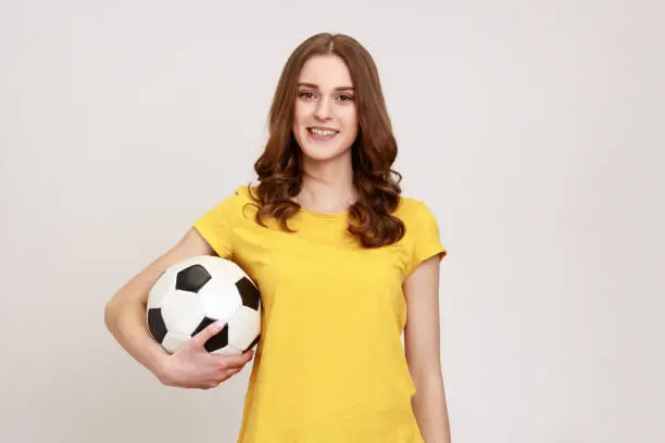 Photo of Photo of joyful teenager girl with brown wavy hair in yellow T- shirt holding soccer ball and supporting favorite team, looking smiling at camera.