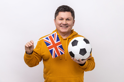 Portrait of satisfied man holding England flag and soccer black and white ball, united football league, wearing urban style hoodie. Indoor studio shot isolated on white background.