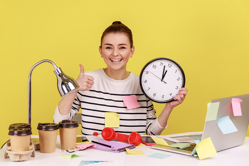 Happy smiling woman covered with sticky notes showing thumbs up and holding wall clock, finishing work on time, time management. Indoor studio studio shot isolated on yellow background.