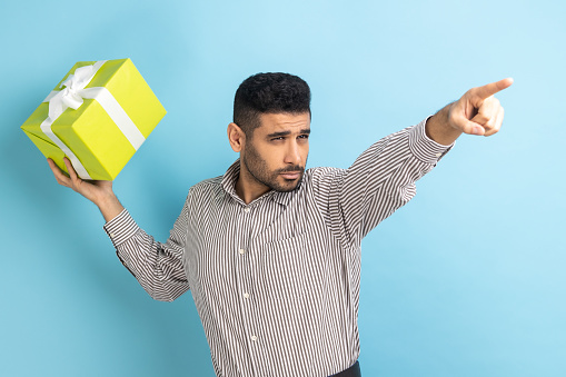 Portrait of young adult serious businessman swung and wants to throw off your gift box, pointing finger in distance, wearing striped shirt. Indoor studio shot isolated on blue background.