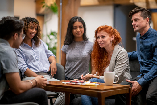 A small group of five adults meet together in a coffee shop to discuss their small business projections.  They are each dressed casually and are seated around a small coffee table as they look over documents together and sip coffee.