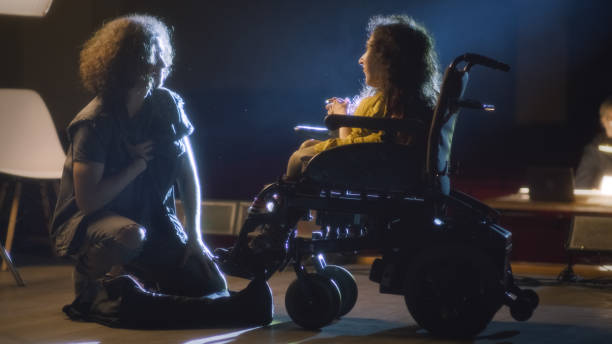 An actor kneeling next to a disabled actress in a wheelchair acting a romantic scene of a play in the theater, during a rehearsal with the director