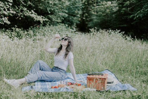 A young beautiful Caucasian girl with brown hair meets in jeans and a t-shirt sits on the floor with a wicker basket, fruits in a plate and seductively eats cherries against the backdrop of tall grasses and trees in a public park, close-up view. parks and forests, in fact, there is a healthy lifestyle, lonely rest.