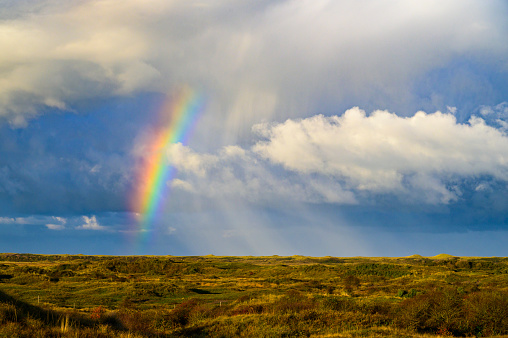 Full arc of double rainbow over summer evening rural landscape - panoramic view of hills with deep forests covered, golden colored meadow with sun beams illuminated, village near forest