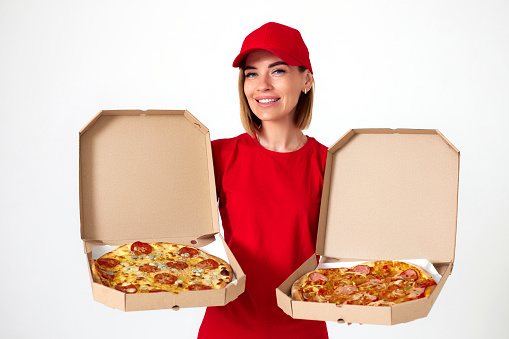 Friendly pizza delivery girl in uniform showing pizza inside boxes on white background