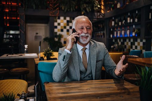 Happy and handsome senior businessman sitting in restaurant and waiting for lunch. He is using smart phone and talking with someone. Business seniors lifestyle concept.