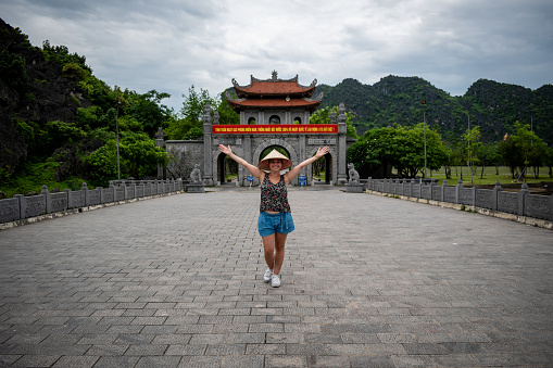Young tourist walking through the ancient capital of Hoa Lu, Vietnam. Young woman with outstretched arms taking a photo in front of the gates of Hoa Lu, Vietnam. Tourist with vietnamese hat touring famous places in Vietnam