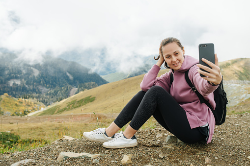 Taking selfie happy young woman sitting in grassy mountains surrounded by low hanging clouds. She is wearing backpack and a pink hoodie.