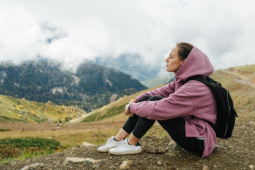 Side view of a woman sitting in grassy mountains surrounded by low hanging clouds. She is wearing backpack and a pink hoodie.