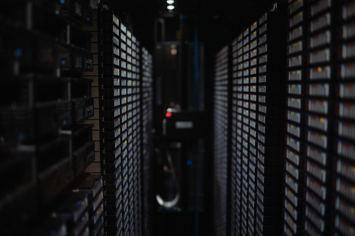 Closeup of a dark data center with multiple rows of fully operational server racks inside a digital information room. Various shelves filled with wired hard drives in a companies database facility