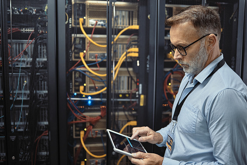 Technician using a digital tablet while working in a server room. Mature man analyzing a code to fix computer networks while doing maintenance in a datacenter. Engineer updating software on a machine