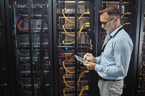 IT technician using a digital tablet in a server room. Mature programmer fixing a computer system and network while doing maintenance in a datacenter. Engineer updating security software on a machine