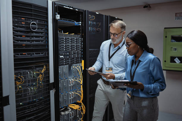 IT technicians using a digital tablet in a server room. Programmers fixing a computer system and network while doing maintenance in a datacenter. Engineers updating security software on a machine stock photo