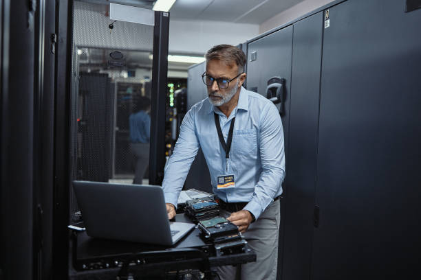 Mature software engineer using a laptop in a server room. IT technician working on network programming with online technology. Protecting information on the internet with cyber security management stock photo