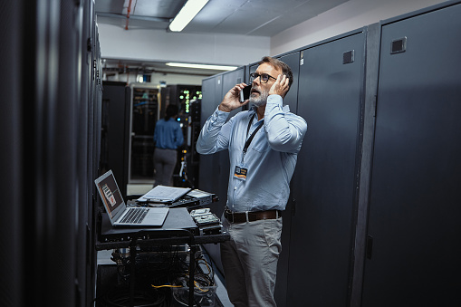 Stressed IT technician talking on the phone while standing in a server room. Male engineer looking worried and anxious about problem while doing server maintenance and repairs in a data center