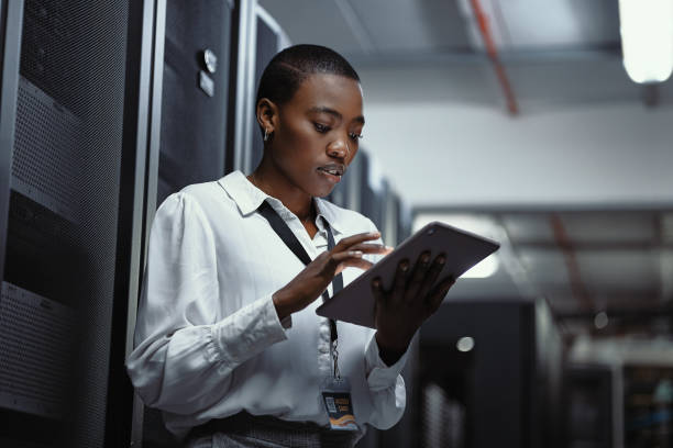 IT technician using a digital tablet in a server room. Female programmer fixing a computer system and network while doing maintenance in a datacenter. Engineer updating security software on a machine stock photo