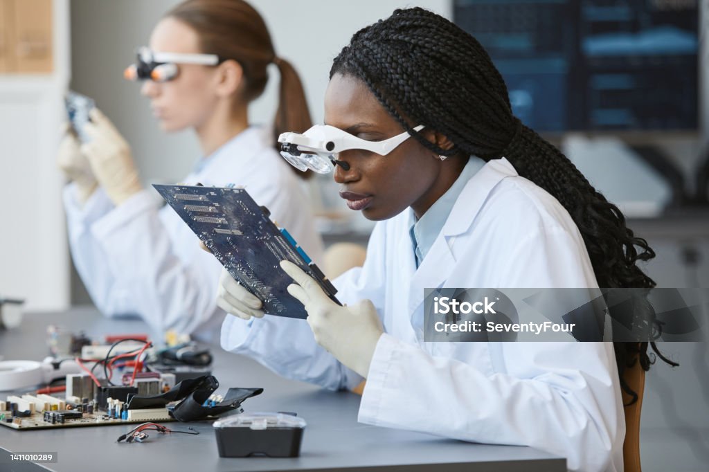 Engineers Working with Tech Portrait of young black woman wearing magnifying glasses and inspecting electronic parts in quality control lab STEM - Topic Stock Photo