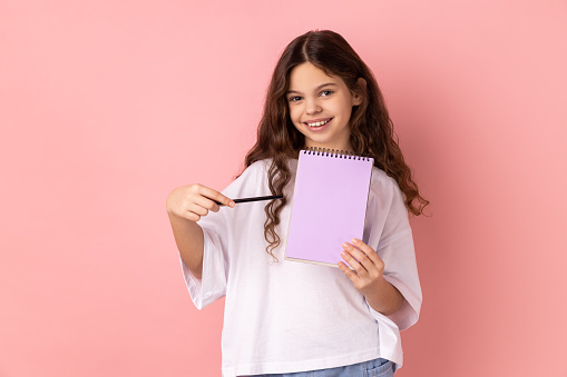 Portrait of beautiful little girl wearing white T-shirt pointing at paper notebook, looking smiling at camera, has positive expression. Indoor studio shot isolated on pink background.