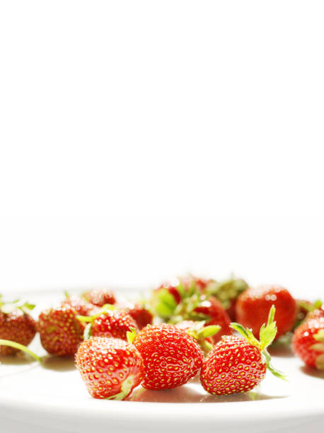 Fresh strawberries with leaves are scattered on the white plate on the bottom of the white isolated backgroung. Juicy strawberries for vegan, vegeterian jams, desserts, cakes, juices. Two strawberries are in focus, the rest is blurred. Bokeh Effect. Close-up. Copy space. Side view