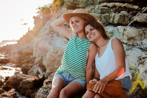 Two young cheerful women in hipsters hats on a rock on the coast of the sea. Summer landscape with girl, sea, Islands and orange sunlight. Travel and tourism with a friend. Twin sister, family