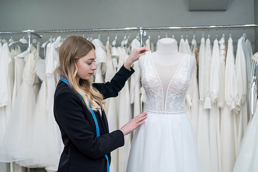 Charming woman saleswoman in stylish black suit measures wedding dress with measuring tape in boutique