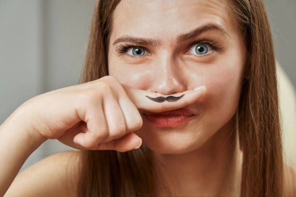 uma jovem engraçada segura um dedo perto de seu nariz, em que um bigode preto é desenhado. hormônios, depilação indesejada - lingerie women spa horizontal - fotografias e filmes do acervo
