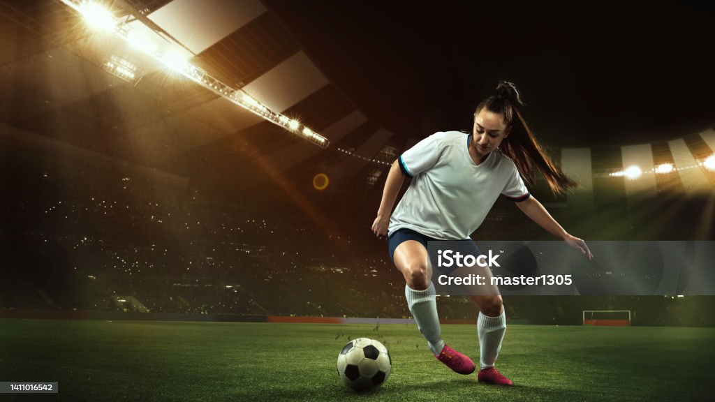 Female soccer, football player dribbling ball in motion at stadium during sport match over evening sky background. Female soccer, football player dribbling ball in motion at the stadium during sport match on evening sky background. Collage at stadium with flashlights. Sport competition. Action, motion, energy and Soccer Stock Photo