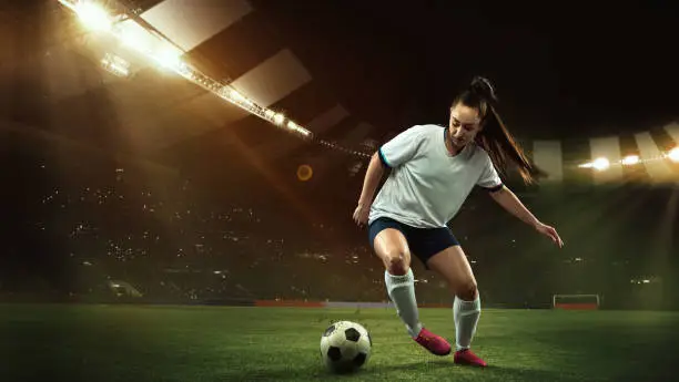 Photo of Female soccer, football player dribbling ball in motion at stadium during sport match over evening sky background.