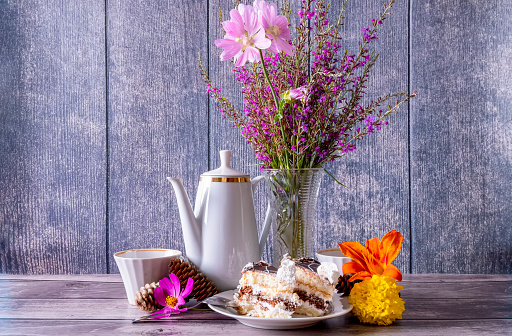 Positive summer still life with coffee pot, wildflowers in vase, slices of cake in plate and coffee mugs on dark wooden background.