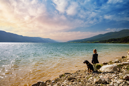 Little girl stands near her big dog friend of Rottweiler breed on empty wild rocky shore near transparent turquoise lake, against backdrop of green Rhodope mountain range covered with spruce forest