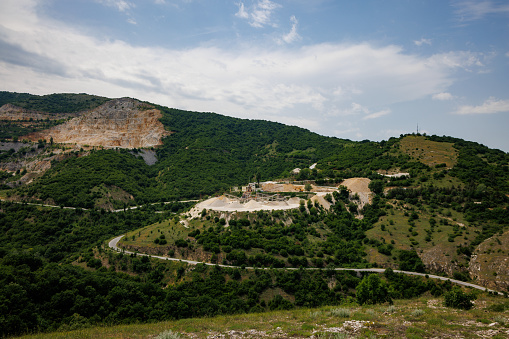 Industrial quarry for extraction of minerals and sand with professional equipment and machines, and intermountain winding road passing by, in Rhodope Mountains covered with coniferous forests