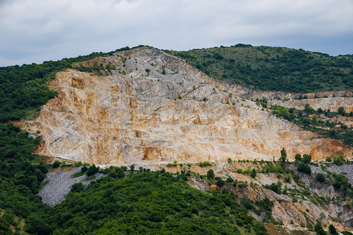 A natural stepped quarry rich in minerals is located near winding intermountain road, against backdrop of Rhodope Mountains and hills with extensive evergreen spruce forests and mountain vegetation