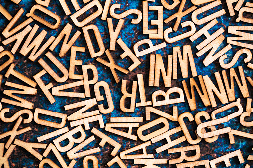 Color image depicting an overhead view of a large selection of wooden alphabet letter tiles scattered, apparently haphazardly, across a blue metallic surface.