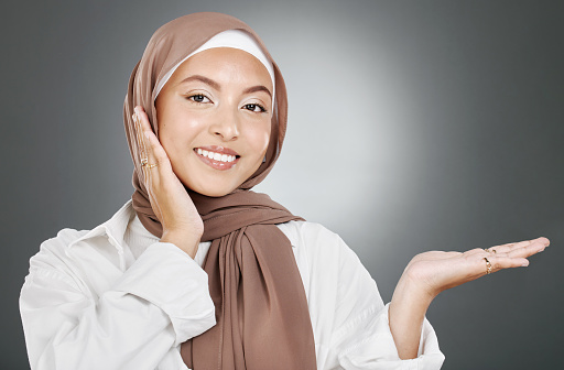 Portrait of glowing beautiful muslim woman isolated against grey studio background. Young woman wearing a hijab or headscarf showing flawless skin and holding her hand out for a product or treatment