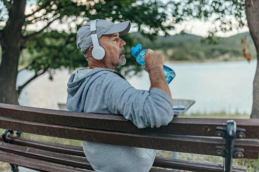 Side view photo of a tired senior man sitting on a bench drinking water to hydrate himself.