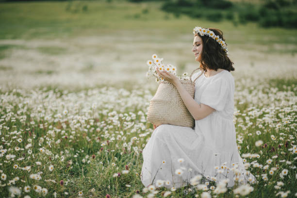 Young woman wearing white dress holding straw basket with flowers on chamomile field. Young woman wearing white dress holding straw basket with flowers on chamomile field. Cottagecore aesthetic. cottagecore stock pictures, royalty-free photos & images