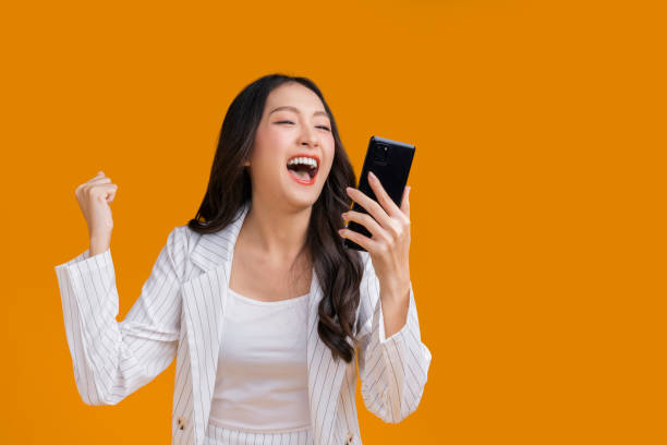 exited surprise face expression asian business woman female hand gesture exited with successful progress result on smartphone screen display  exited raised hands up isolated on bright yellow color - extatisch stockfoto's en -beelden