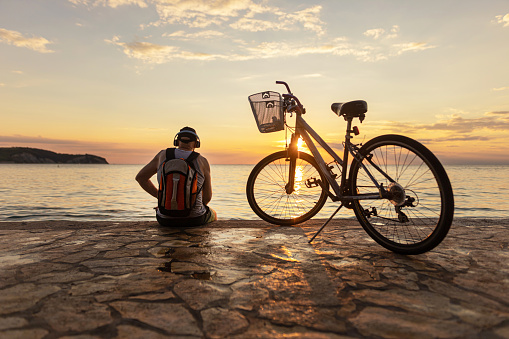 Rear view of sunset and calm sea, senior man sitting and pausing riding a bike and listening music.