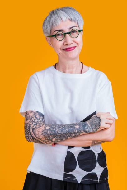 cheerful smiling asian senior elder woman wear glasses grey hair in casual cloth hand tattoo abstract pattern portrait headshot on yellow color background healthy lifestyle stock photo