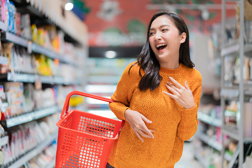 smiling cheerful and joyful female woman hand hold shopping basket hand gesture greeting surprise standing between supermarket product shelf aisel convenience store supermarket department store mall