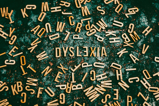 Color image depicting an overhead view of wooden letter tiles spelling out the word Dyslexia, with the 'E' placed backwards. The word is surrounded by a large collection of other alphabet letter tiles, in a haphazard arrangement.