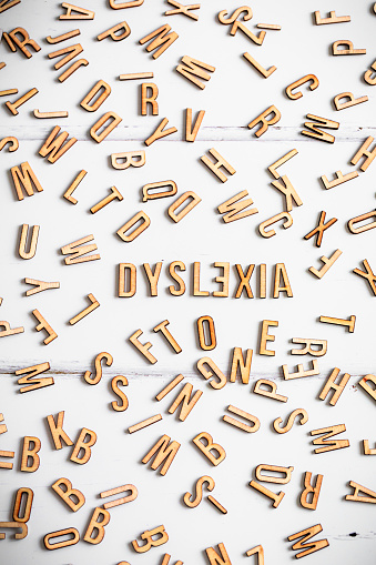 Color image depicting an overhead view of wooden letter tiles spelling out the word Dyslexia, with the 'E' placed backwards. The word is surrounded by a large collection of other alphabet letter tiles, in a haphazard arrangement.