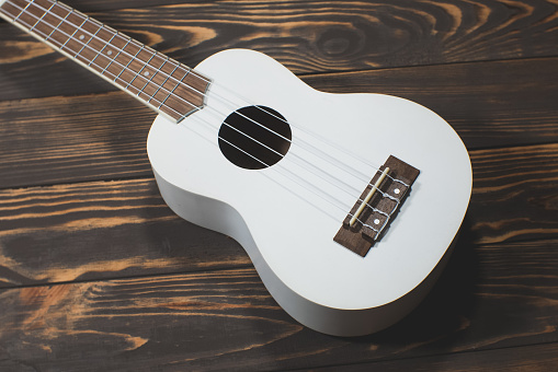 White colored wooden ukulele guitar on the brown wooden background. Hawaiian Four String Guitar. Musical Instrument.
