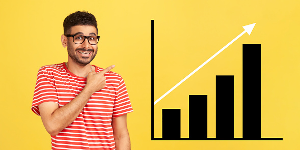 Portrait of happy joyful young bearded man standing, pointing aside and showing business growth graph. indoor studio shot isolated on yellow background