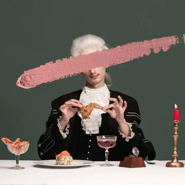 Dinner. Creative portrait of young elegant man in peruke and vintage jacket with stroke of pink paint sitting at table isolated on dark green background. Retro style, comparison of eras concept.