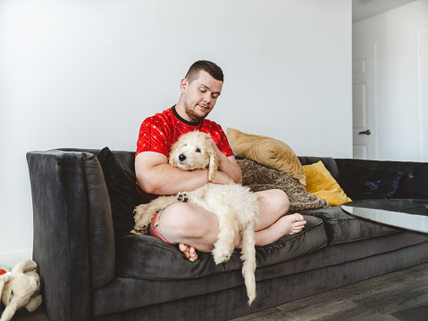 Young Gay man with his puppy dogs at home. He is wearing summer outfits with red shirt and colourful short. Interior of urban apartment in North America.