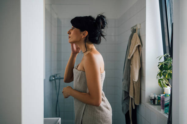 A Beautiful Pensive Woman Relaxing After Taking A Shower A side view of a smiling Caucasian female wrapped in a towel standing in the bathroom after having a bath and taking care of her skin. woman wrapped in bath towel stock pictures, royalty-free photos & images