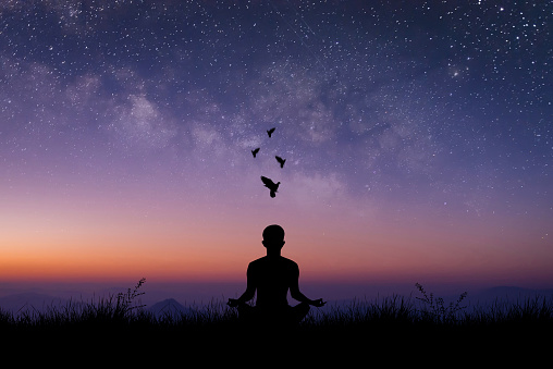 Silhouette of young male sitting practices yoga and meditating in lotus position alone on top of the mountain with night sky, star and Milky Way. He felt calm and happy.